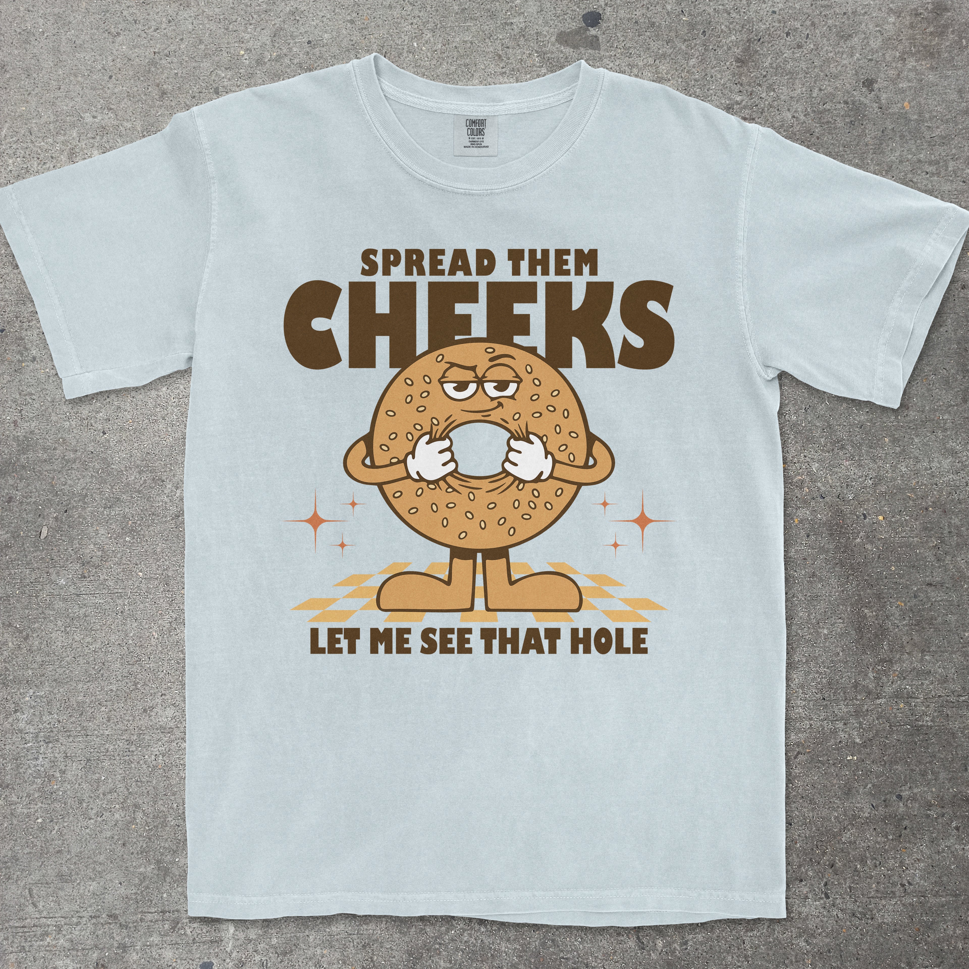 Light blue t-shirt with a cartoon bagel character and humorous text on a concrete surface