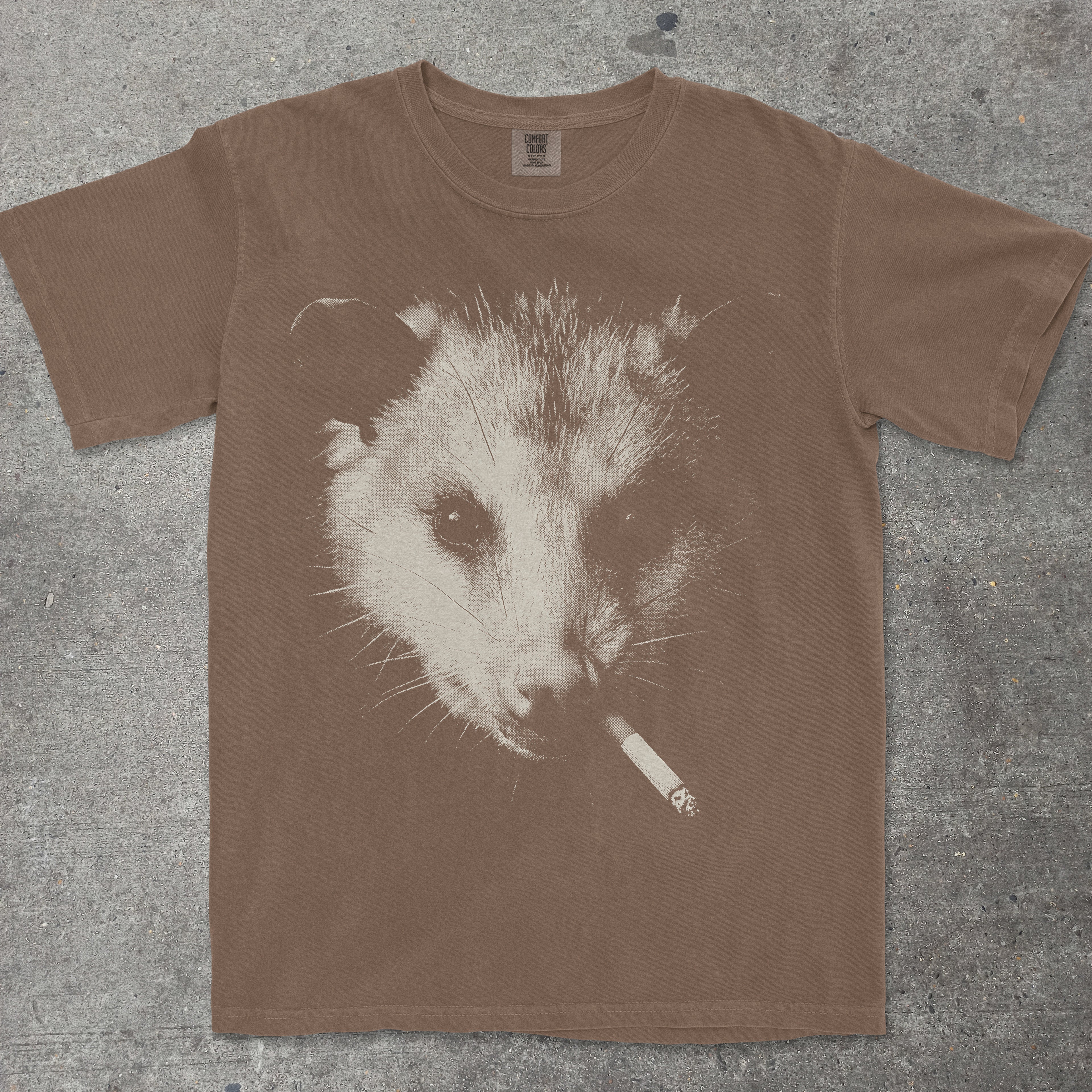 Brown t-shirt with an opossum graphic holding a cigarette on a concrete surface