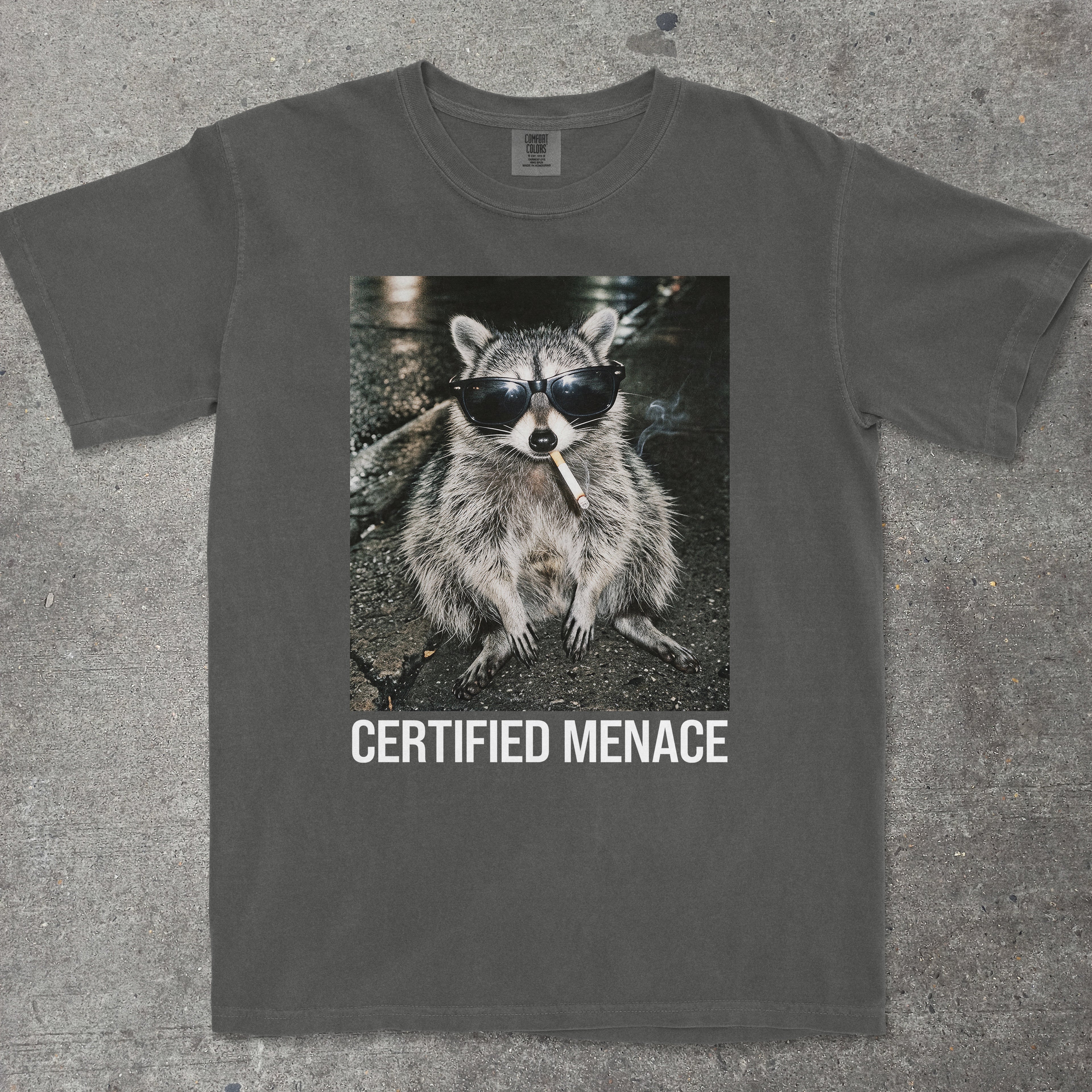 T-shirt with a raccoon wearing sunglasses and smoking a cigarette, labeled 'Certified Menace', on a textured surface.
