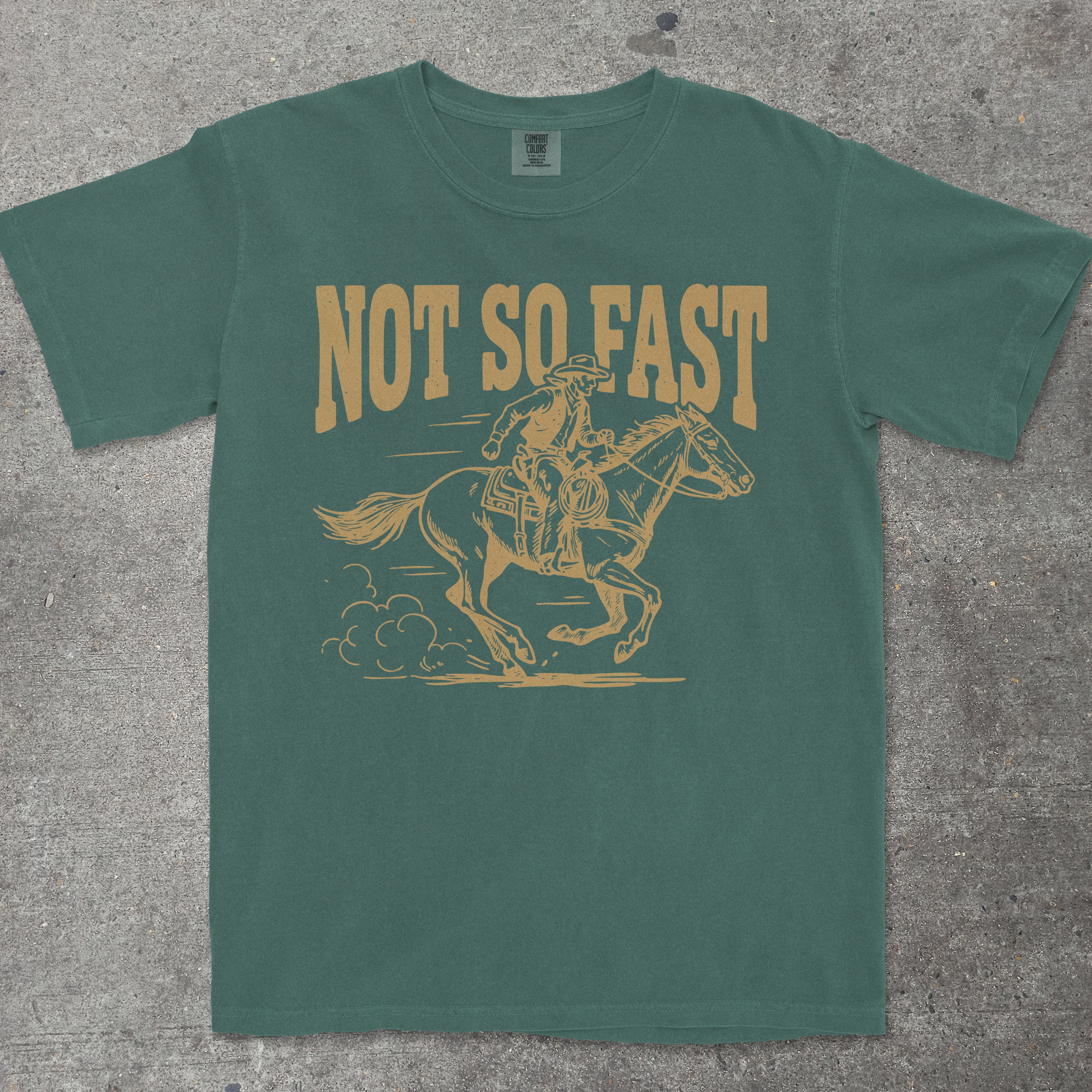 Green t-shirt with yellow graphic of a person on a horse and 'Not So Fast' text on a concrete surface.